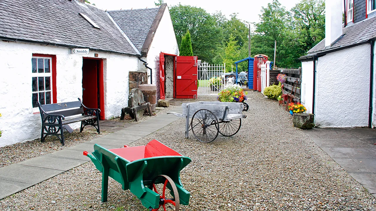 White buildings at the Arran Heritage Museum with red doors. There are wheelbarrows, flowers and a bench in the middle of the courtyard