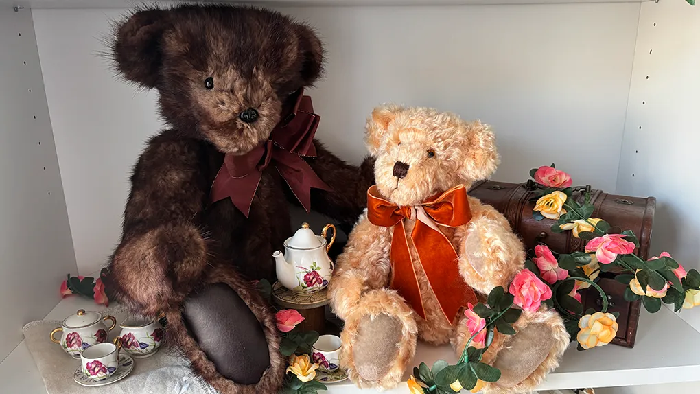 Two teddy bears, one big brown bear and one smaller blonde bear. They are surrounded by flowers, a tea set and a chest