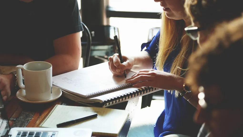 People sitting at a table, there is one person writing. A coffee cup, notepad and laptop are also on the table.